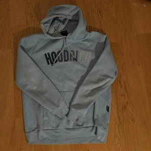 Ljusblå hoodie från Hoodrich - Säljer en ljusblå hoodie från Hoodrich med stor broderad logga på bröstet. Tröjan har huva med snörning och en stor magficka framtill. Perfekt för dig som gillar streetwear och vill ha en chill look.