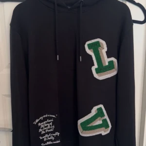 Svart hoodie från Louis Vuitton - Svart hoodie från Louis Vuitton med stora gröna och beige applikationer på framsidan. Tröjan har huva med snörning och tryckt text på ena sidan. Tillverkad i mjuk bomull och har en avslappnad passform med långa ärmar.