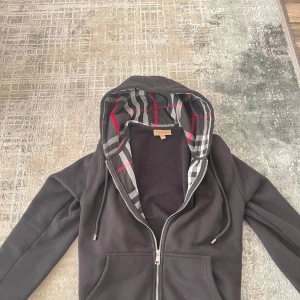 Burberry hoodie - Svart hoodie från Burberry med klassiskt rutigt foder i huvan i svart, vitt och rött. Tröjan har dragkedja framtill, snörning i huvan och broderad logga på bröstet. Tillverkad i mjuk bomullsmix, perfekt för kyliga dagar.