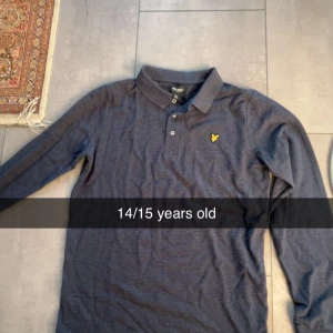 Mörkblå långärmad pikétröja Lyle & Scott - Snygg mörkblå långärmad pikétröja från Lyle & Scott med klassisk krage och tre knappar framtill. Tröjan har det ikoniska gula logomärket broderat på bröstet. Perfekt för dig som gillar stilrena och tidlösa plagg.