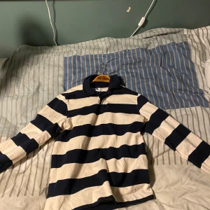 Randig pikétröja från These Glory Days - Säljer en klassisk långärmad pikétröja från These Glory Days i storlek small. Tröjan har breda marinblå och vita ränder, krage och knäppning vid halsen.Tillverkad i mjuk bomull.