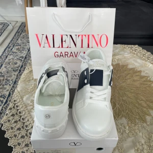 Valentino Garavani vita sneakers - Snygga vita sneakers från Valentino Garavani med bred marinblå rand och coola nitar på sulan. Skorna har snörning, rund tå och är tillverkade i skinn. Perfekta för dig som vill ha en stilren men ändå lyxig look.