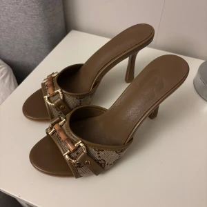 Bruna pumps med gulddetaljer - Snygga bruna pumps med hög klack och öppen tå. Ovandelen är i canvas med monogrammönster och detaljer i brunt skinn samt guldfärgade spännen. Perfekta för dig som vill ha en chic och trendig look med lyxig känsla.