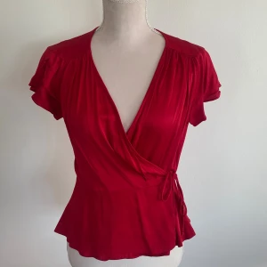 Röd omlottblus med knyt i midjan från Zara - Röd kortärmad omlottblus med v-ringning och volanger. Blusen har ett knyt i midjan och har även en knapp på insidan av blusen för extra säkerhet! 