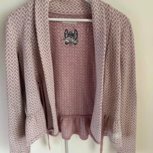 Rosa och vit kofta med volangkant - Supersöt kofta från Odd Molly i rosa och vitt med zigzag-mönster. Koftan har volangkant nertill och vid ärmslut, samt knytband i midjan. Perfekt för lager på lager och ger en chill men ändå söt vibe.