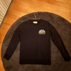 Svart sweatshirt från Casablanca - Svart sweatshirt från Casablanca med färgglatt tryck av byggnad och palmer på ryggen samt mindre tryck på bröstet. Tröjan har rund hals, långa ärmar och ribbade muddar. Perfekt för dig som gillar statement-plagg och snygga prints.