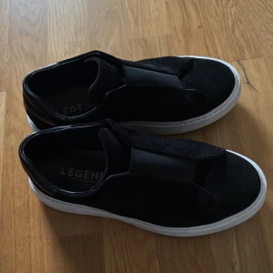 Arigato legends skor, priset går att diskuteras - Svarta sneakers från Legend med stilren design och bred resår över vristen istället för snörning. Ovandelen är i mocka och detaljer i skinn, med en klassisk vit platt sula. Perfekta för dig som gillar minimalistisk och clean stil.