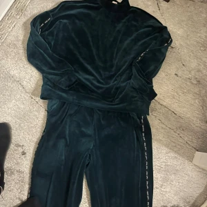 Mörkgröna mjukisbyxor från Hunkemöller - Mörkgröna mjukisbyxor från Hunkemöller i sammetsliknande material. Byxorna har en rak passform och resår i midjan. Sidorna är dekorerade med en smal textremsa för en sportig vibe. Perfekta för chill dagar hemma.