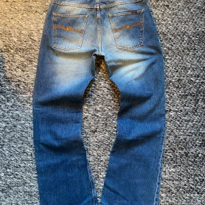 Nudie Jeans  - Säljer ett par klassiska blå jeans från Nudie Jeans med orange sömmar och ikonisk bakficka. Väldigt bra skick, enda tecken på användning är i skrevet där man kan ana att de har cyklats i. Modellen Flare Glenn har bootcut-ben och knäppning med knappar framtill. Jeansen är i 100% bomull. Storlek 33/32 men skulle säga att de även passar 32/32. Nypris 1800 sek. Hör av dig vid frågor eller funderingar om mått eller annat! Priset kan alltid diskutera :) 