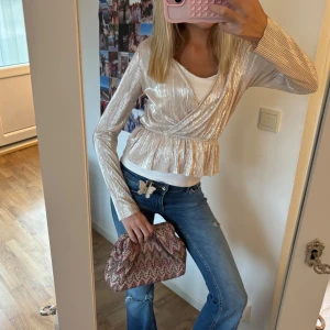 Glittrig omlottopp i champagne - Säljer en glittrig omlottopp i champagnefärg med lång ärm och v-ringning. Toppen har plisserat tyg, markerad midja och volang nertill. Perfekt för dig som vill sticka ut med en trendig och festlig look.