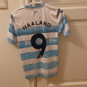 Manchester City Haaland tröja Puma 164 - Säljer en Manchester City fotbollströja från Puma med Haaland och nummer 9 på ryggen. Tröjan är vit med ljusblåa horisontella ränder, klubbmärke och sponsortryck på bröstet. Tillverkad i lätt och ventilerande material, perfekt för match eller träning.