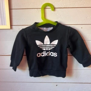 Svarta Adidas mjukisbyxor baby - Svarta mjukisbyxor från Adidas med vita ränder längs sidorna och klassisk Adidas-logga framtill. Byxorna har snörning i midjan och muddar vid bensluten. Perfekta för en sportig och chill stil till de minsta.