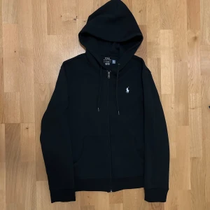 zip hoodie från Polo Ralph Lauren - Svart zip hoodie från Polo Ralph Lauren med dragkedja framtill, luva med snörning och logga på bröstet. Hoodien är i storlek S och är i nyskick och är oanvänd.