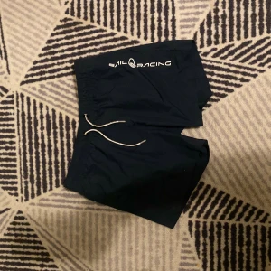 Svarta Sail Racing badshorts med snörning - Svarta badshorts från Sail Racing med vit logga på ena benet och vit snörning i midjan. Klassisk loose passform och elastisk midja. Perfekta för stranden eller poolen, tillverkade i snabbtorkande syntetmaterial.