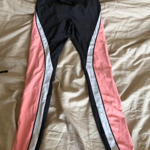 Svarta och rosa leggings med vit detalj - Snygga leggings i svart med breda rosa paneler längs benen och en vit rand som accent. Tight passform och stretchigt material, perfekta för träning eller chill. Sportig look som sticker ut.