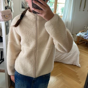 Beige teddyjacka från Kappahl - Supermjuk och fluffig beige teddyjacka från Kappahl med dragkedja framtill och hög krage. Jackan har en loose fit och är perfekt för kyliga dagar. Materialet är fleece-liknande och ger en mysig vibe. Passar dig som gillar en enkel men trendig look.