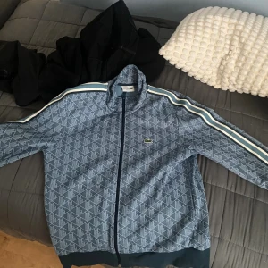 Blå mönstrad hoodie från Lacoste - Säljer en blå hoodie från Lacoste med hel dragkedja och ikoniska ränder längs ärmarna. Tröjan har ett diskret geometriskt mönster över hela ytan och klassisk krokodillogga på bröstet. Perfekt för dig som gillar sportig och stilren streetwear.