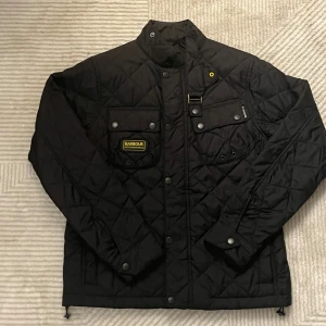 Svart quiltad jacka Barbour International - Svart quiltad jacka från Barbour International med coola detaljer som tryckknappar, bröstfickor och broderad logga. Jackan har hög krage med spänne och snyggt mönstrat innerfoder med motorcykeltryck. Perfekt för dig som gillar stilren och sportig look. Aldrig använd