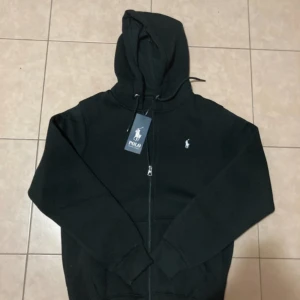 Svart hoodie från Polo Ralph Lauren - Svart hoodie från Polo Ralph Lauren med dragkedja framtill och klassisk logga broderad på bröstet. Tröjan har huva med snörning och långa ärmar. Perfekt för en chill och stilren look.