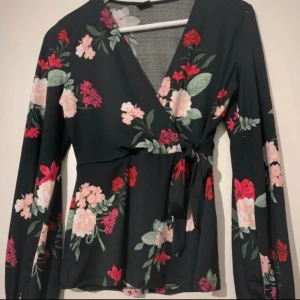 Blommig omlottopp med knytband - Svart omlottopp med blommigt mönster i rosa, rött och grönt. Toppen har långa ärmar och knytband i midjan för en snygg passform. V-ringad och lätt utsvängd nertill, perfekt för en trendig look. Storlek XS, dock väldigt stretch och passar en som vanligtvis har M/L 