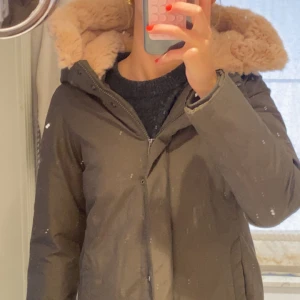 Olivgrön parkas från Woolrich med päls - Säljer en olivgrön parkas från Woolrich med mysig beige fuskpäls i huvan. Jackan har dragkedja och knappar framtill, långa ärmar och två fickor. Perfekt för kalla dagar med sin varma och långa modell.