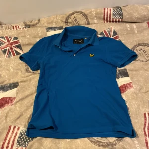 Blå pikétröja från Lyle & Scott - Snygg blå pikétröja från Lyle & Scott med klassisk krage och korta ärmar. Tröjan har det ikoniska gula fågel-loggan broderad på bröstet och är tillverkad i mjuk bomull. Perfekt för en stilren och sportig look. Väldigt lite använd.      Vid funderingar eller pris dig