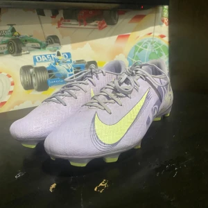 Lila Nike fotbollsskor med neon-detaljer - Säljer ett par snygga lila Nike fotbollsskor med neon-gula detaljer och mönstrad ovandel. Skorna har snörning och platt sula med fasta dobbar, perfekt för gräsplan. Materialet är syntet och designen är riktigt modern med coola grafiska linjer på sidorna.