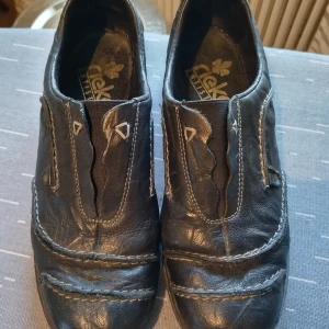 Svarta skinnloafers från Rieker - Svarta loafers från Rieker i mjukt skinn med dekorativa sömmar och små metallnitar. Skorna har elastiska partier för enkel påtagning och en bekväm, platt sula. Perfekta för dig som gillar en klassisk och chill stil.