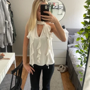 Vit volangblus med halterneck - Supersöt vit blus med volangdetaljer längs framsidan och en snygg halterneck med knyt runt halsen. Ärmlös modell i lätt och flowigt tyg, perfekt för varmare dagar eller när du vill känna dig extra stylish.