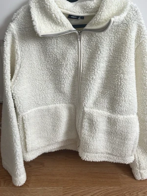 Vit teddyjacka med dragkedja  - Mysig vit teddyjacka med hel dragkedja framtill och stora fickor. Jackan har en fluffig, fårskinnsinspirerad look och är perfekt för kyliga dagar. Långärmad och med en avslappnad passform som ger en chill vibe.