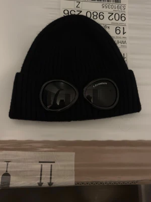 Svart mössa med goggles C.P. Company - Säljer en svart ribbstickad mössa från C.P. Company med ikoniska inbyggda goggles framtill. Mössan har uppvikt kant och är tillverkad i mjukt material som sitter skönt på huvudet. Perfekt för dig som vill sticka ut med en streetwear-look.