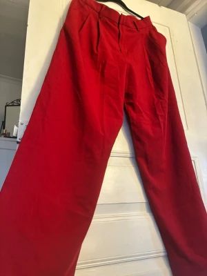 Röda kostymbyxor från Lindex - Snygga röda kostymbyxor från Lindex med hög midja och vida ben. Byxorna har pressveck framtill, bälteshällor och diskreta fickor. Perfekta för dig som vill sticka ut med färg och stil. Helt nya, aldrig använda.