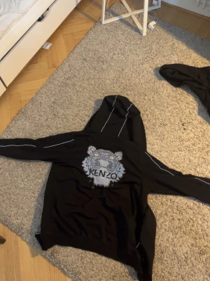 Svart Kenzo hoodie med tigermotiv - Svart hoodie från Kenzo med ikoniskt tigermotiv broderat på ryggen och mindre logga på bröstet. Tröjan har dragkedja, huva och vita kontrastsömmar längs ärmar och huva. Knappt använt och lite ovanligare utgåva storlek står L men sitter som M