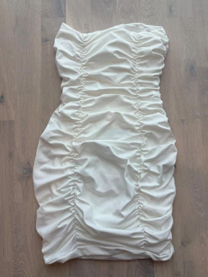 Vit axelbandslös kortklänning - Säljer en vit, axelbandslös kortklänning med snygga rynkade detaljer längs hela klänningen. Klänningen har en tight passform och är perfekt för dig som vill ha en stilren men ändå trendig look. Materialet känns mjukt och stretchigt.