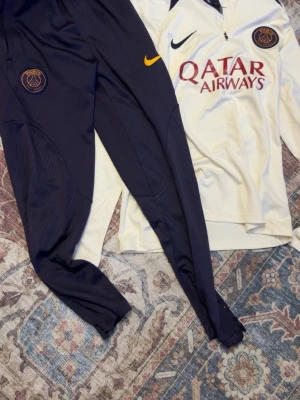 PSG träningsset från Nike - Snyggt PSG träningsset med långärmad vit tröja och svarta byxor. Tröjan har Qatar Airways-logga och PSG-emblem, byxorna har PSG-märke och gul Nike-logga. Perfekt för fotbollsträning eller chill. Materialet är mjukt och sportigt.