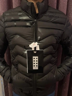 Svart dunjacka från Moncler - Snygg svart dunjacka från Moncler med diskret glansig finish och klassisk ståkrage. Jackan har dragkedja framtill och ett stilrent, quiltat mönster. Perfekt för kalla dagar och enkel att matcha med olika outfits.