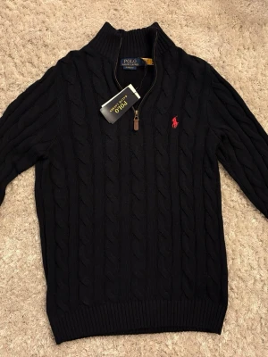 Mörkblå kabelstickad tröja Polo Ralph Lauren - Kabelstickad mörkblå tröja från Polo Ralph Lauren med halv dragkedja och klassisk röd broderad logga på bröstet. Tröjan har ribbade muddar och hög krage, perfekt för en stilren look. Classic fit-modell med exklusiv känsla.