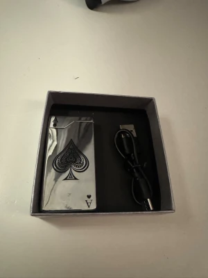 Elektrisk tändare ess i metall - Elektrisk tändare i silverfärgad metall med ess-symbol på framsidan. Laddas via USB och har blå LED-ljus när den används. Kommer i en ask tillsammans med laddningskabel. Perfekt för dig som gillar snygga accessoarer med korttema. Pris kan diskuteras 