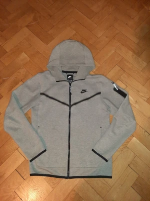 Grå Nike Tech Fleece hoodie - Snygg grå Nike Tech Fleece hoodie med hel dragkedja och svarta detaljer. Hoodien har en huva, svarta linjer över bröstet och ärmarna samt Nike-logga på vänster sida. Tillverkad i mjukt och bekvämt material, perfekt för en sportig och avslappnad stil. Och ALDRIG ANVÄND.