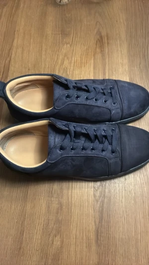 Mörkblå sneakers från Louboutin - Snygga mörkblå sneakers från Louboutin med klassisk röd sula och ovandel i mocka. Skorna har rund tå, platt sula och snörning. Perfekta för dig som vill ha en stilren och lyxig look.