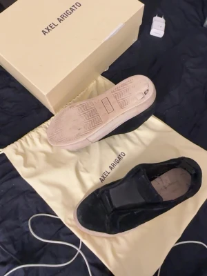 Svarta slip-on sneakers Axel Arigato - Stilrena svarta slip-on sneakers från Axel Arigato med vit sula. Skorna har en minimalistisk design och är tillverkade i mocka med elastiska paneler på sidorna för enkel påtagning. Perfekta för en clean och modern look. Du får med allt som är på bilden! Slitage på sulor och även vid hälen 