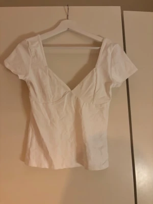 Vit topp med v-ringning och kort ärm - Säljer en vit topp med djup v-ringning både fram och bak. Toppen har korta ärmar och en figurnära passform som framhäver siluetten. Perfekt till jeans eller kjol för en clean och fräsch look.