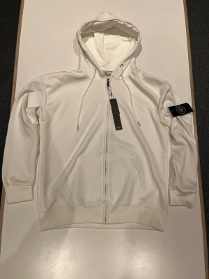 Vit zip-hoodie från Stone Island - Snygg vit hoodie med dragkedja från Stone Island. Klassisk huva med dragkedja, två stora fickor framtill och ikonisk logotyp-patch på ärmen. Tillverkad i mjukt material som känns skönt mot huden. Perfekt för en clean och stilren look.