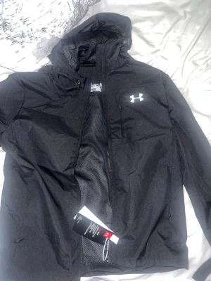 Svart vindjacka från Under Armour - Snygg svart vindjacka från Under Armour med huva och dragkedja framtill. Jackan har en vit logga på bröstet, meshfoder på insidan och praktiska fickor. Perfekt för blåsiga dagar och sportiga outfits.