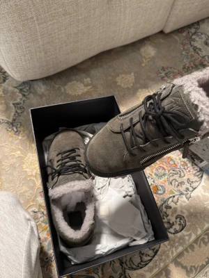 Grå sneakers från Giuseppe Zanotti - Sneakers i grå mocka från Giuseppe Zanotti med svart snörning, dragkedja på sidan och mjukt foder i fuskpäls. Snygg och robust design som passar perfekt till höst och vinter. Kommer i originalkartong.