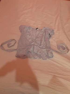 Blårandig omlottopp med volang - Söt blårandig omlottopp med knytband i midjan och volangkant. Toppen har korta ärmar och v-ringad hals. Perfekt för varma dagar och ger en fräsch sommarkänsla. Materialet känns lätt och luftigt.