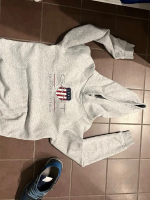 Grå hoodie från GANT med tryck - Snygg ljusgrå hoodie från GANT med klassiskt tryck och amerikansk flagga på bröstet. Hoodien har huva, magficka och ribbade muddar vid ärmar och nederkant. Perfekt för en avslappnad och sportig stil. Materialet känns mjukt och bekvämt.