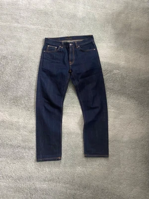 Mörkblå selvage jeans från Nudie Jeans W31 L30 - Säljer ett par nya mörkblå selvage jeans från Nudie Jeans med klassisk orange kontrastsöm och ikoniska bakfickor med broderad våg. Jeansen har rak passform och är tillverkade i kraftig denim av bomull. Modell Gritty Jackson dry selvage. Perfekta för dig som gillar stilrena och tidlösa jeans.