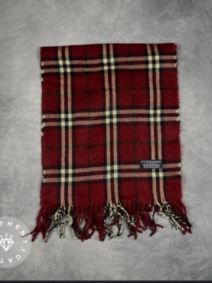 Rutig vinröd halsduk från Burberry - Klassisk halsduk från Burberry i vinrött med rutigt mönster i beige, svart och vitt. Tillverkad i 100% lammull och har fransar i ändarna. Perfekt accessoar för att lyfta din outfit med en tidlös brittisk stil. Köpt på tradera och är markerad som äkta.