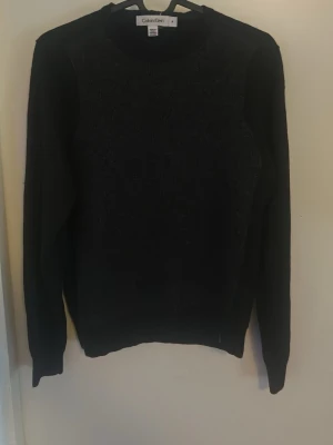 Svart stickad tröja från Calvin Klein - Snygg svart stickad tröja från Calvin Klein med rund halsringning och långa ärmar. Klassisk och enkel design som passar till det mesta. Perfekt för dig som gillar stilrena plagg med en exklusiv touch.